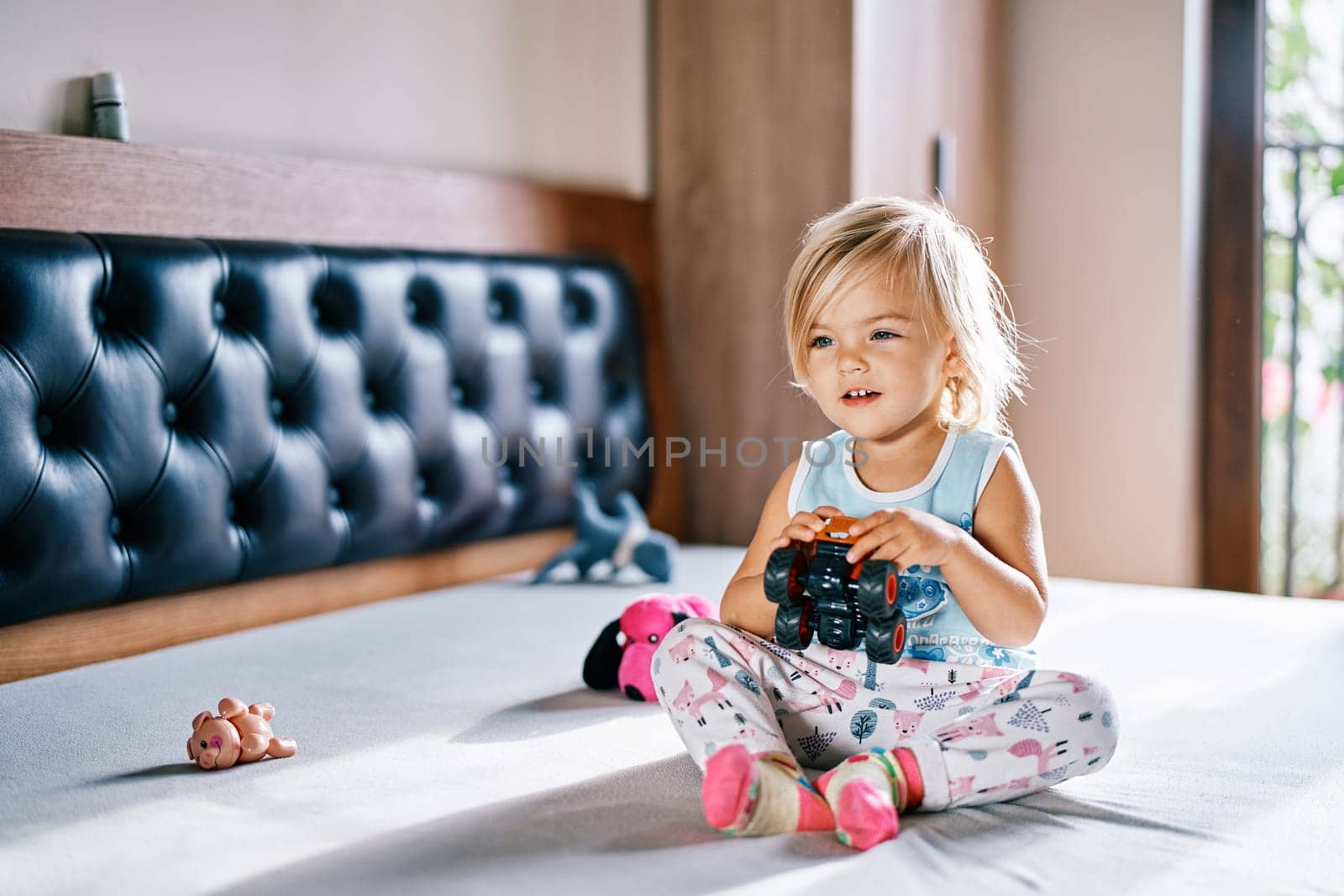 Little smiling girl sitting on the bed with a toy car in her hands. High quality photo