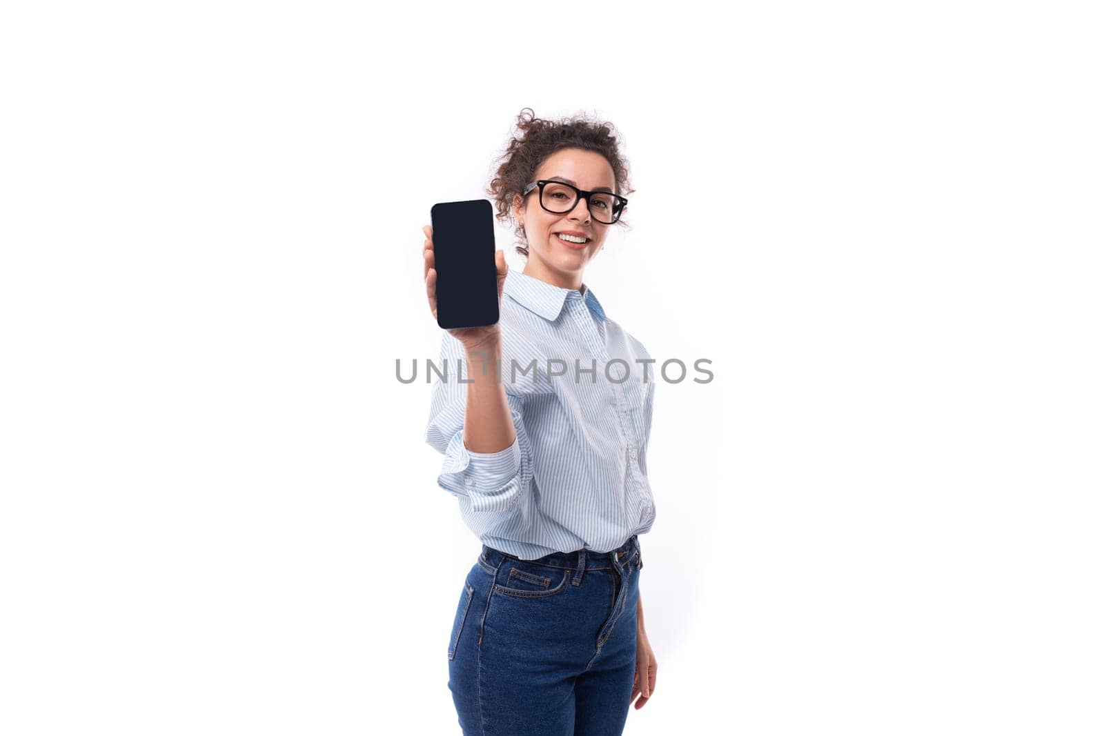portrait of a young smart brunette assistant lady dressed in a basic blue shirt and jeans holding a smartphone with the screen forward in front of her by TRMK