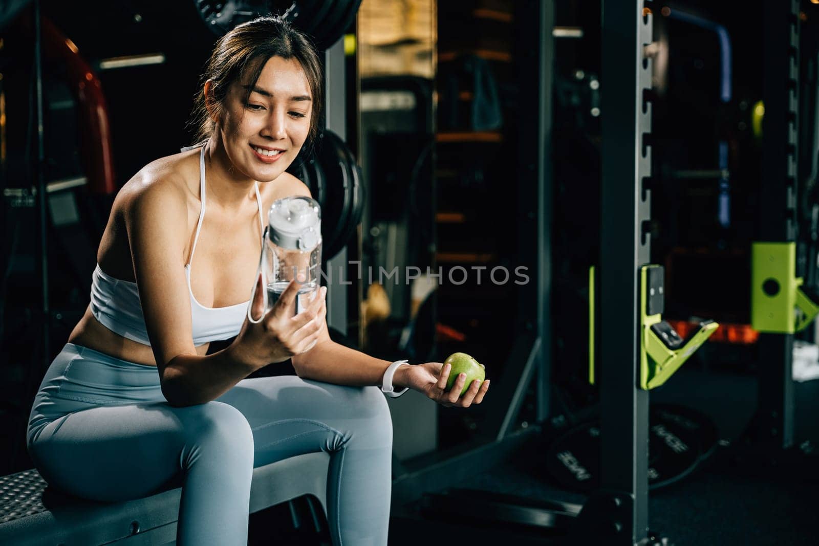 Woman holds a green apple and a water bottle in gym, emphasizing the importance of healthy food choices and staying hydrated for maintaining a healthy lifestyle. Healthy fitness and eating lifestyle