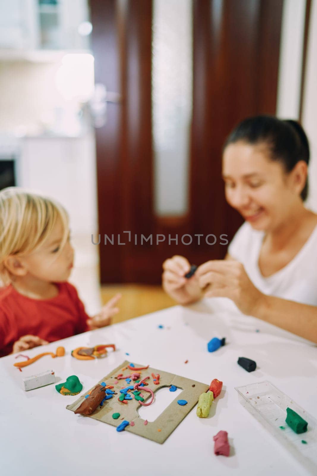 Mom and her little daughter sculpt different figures from colored plasticine while sitting at the table by Nadtochiy