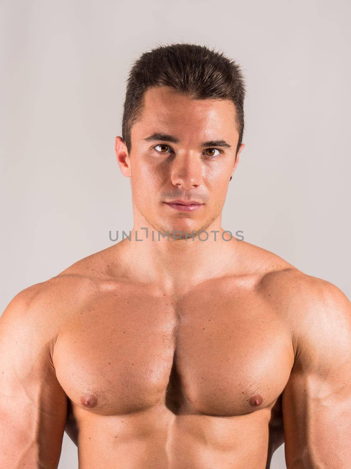 Shirtless young male bodybuilder in studio portrait, looking at camera on neutral background, showing muscular torso and body