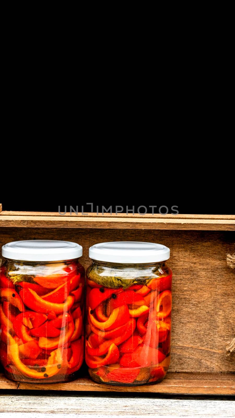 Wooden crate with glass jars with pickled red bell peppers.Preserved food concept, canned vegetables isolated in a rustic composition. by vladispas