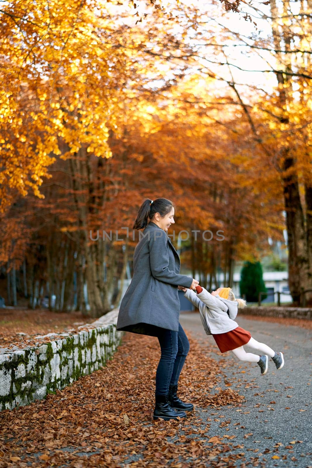 Mom spins a little girl by the arms while standing on the road in the autumn park by Nadtochiy