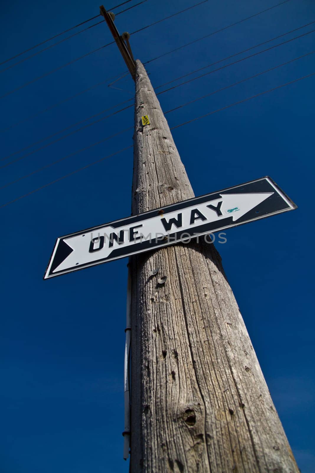 Clear Direction: A weathered utility pole in Allegan, Michigan proudly displays a 'ONE WAY' sign, pointing left against a vast blue sky, symbolizing certainty, decision-making, and a singular path forward.