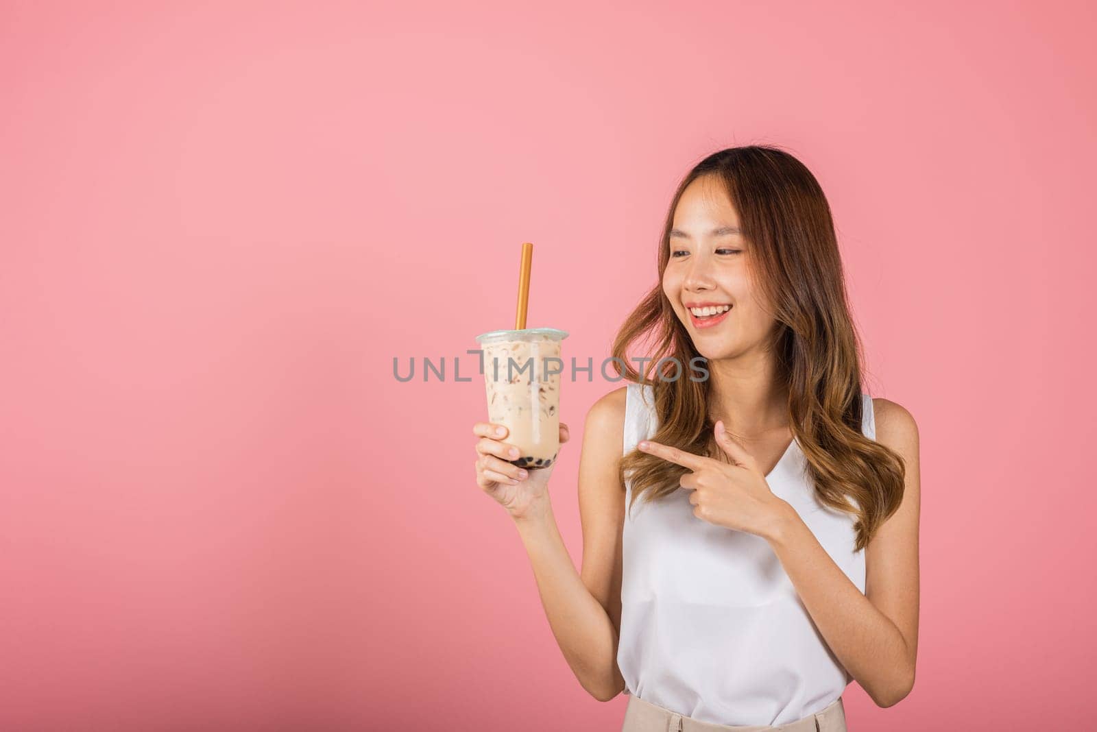 Asian beautiful young woman holding drinking brown sugar flavored tapioca pearl bubble milk tea and pointing finger, smile portrait female, isolated on pink background, pearl milk tea beverage concept