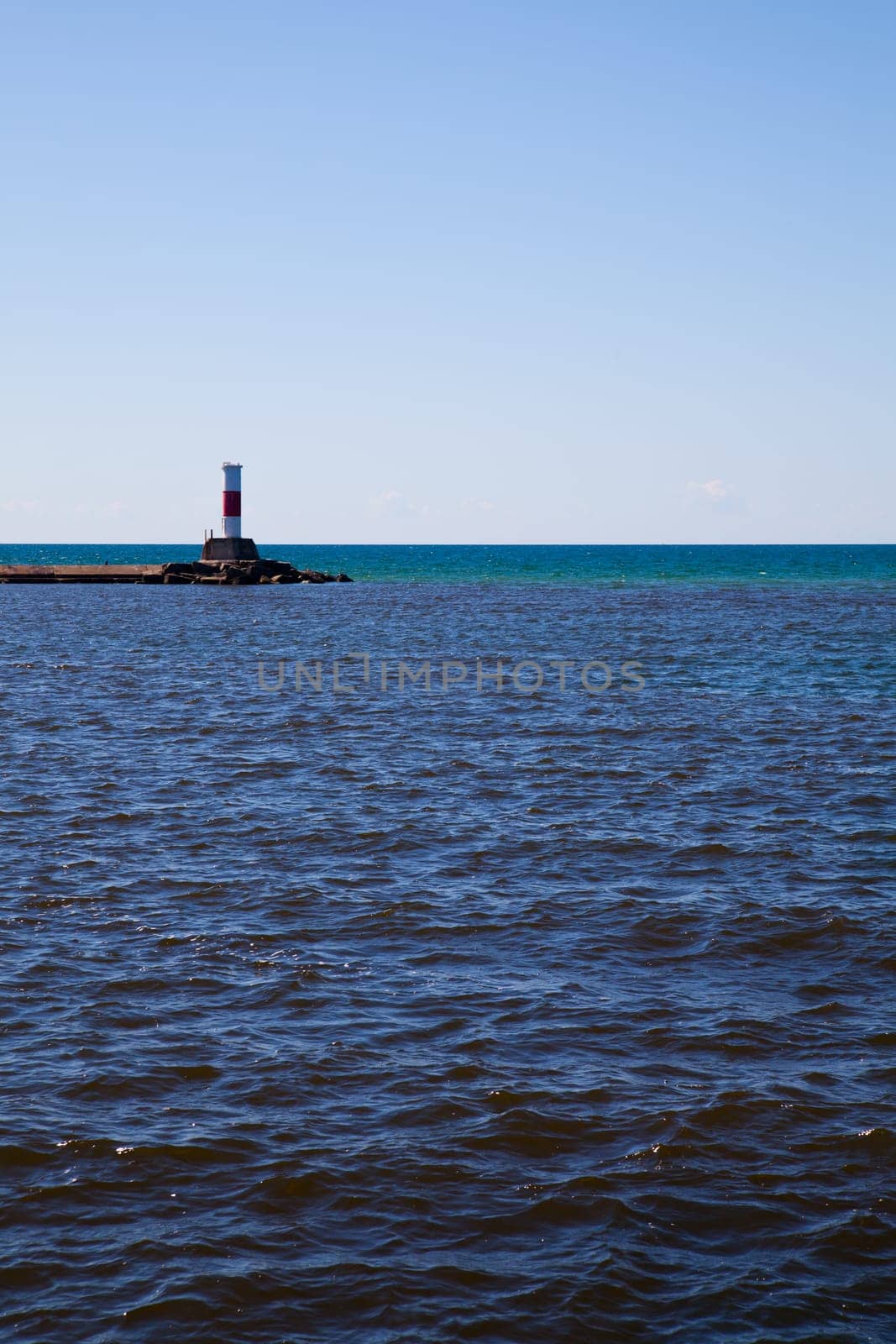 Tranquil coastal landscape featuring a striking red and white lighthouse against a clear blue sky and calm waters. Perfect for maritime industries and travel promotions.