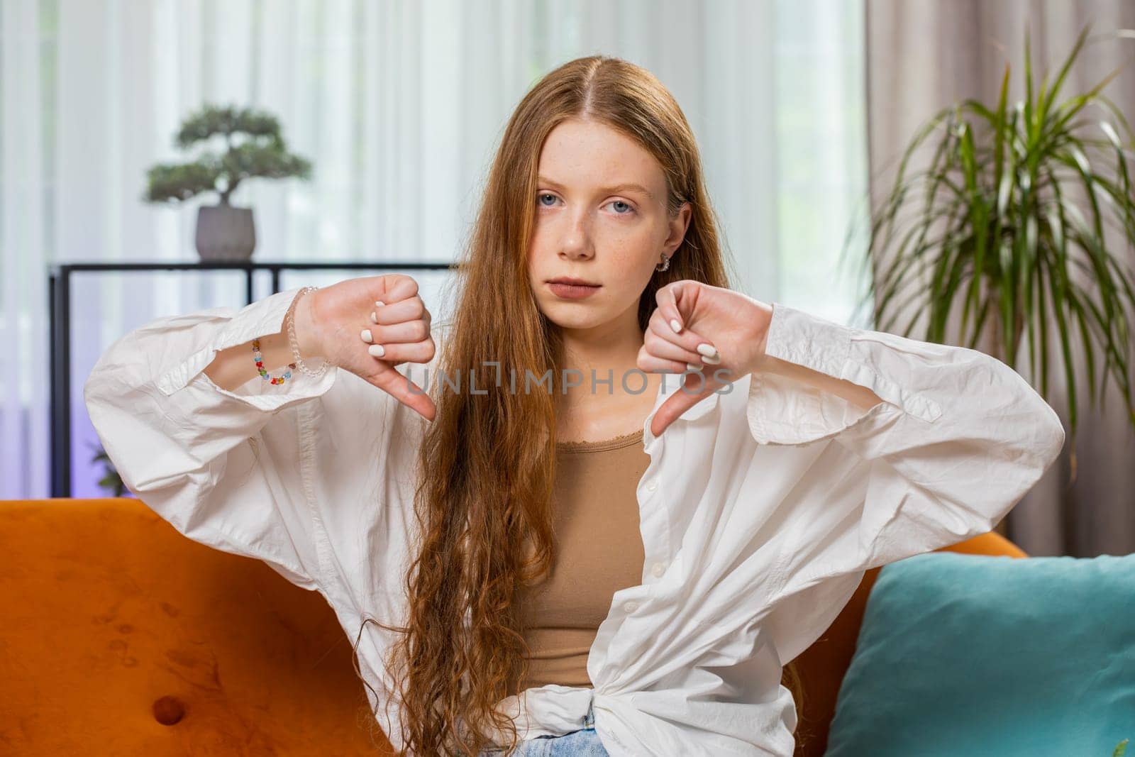 Dislike. Portrait of upset girl showing thumbs down sign gesture, expressing discontent, disapproval dissatisfied bad work at modern home apartment indoors. Displeased child in living room on sofa