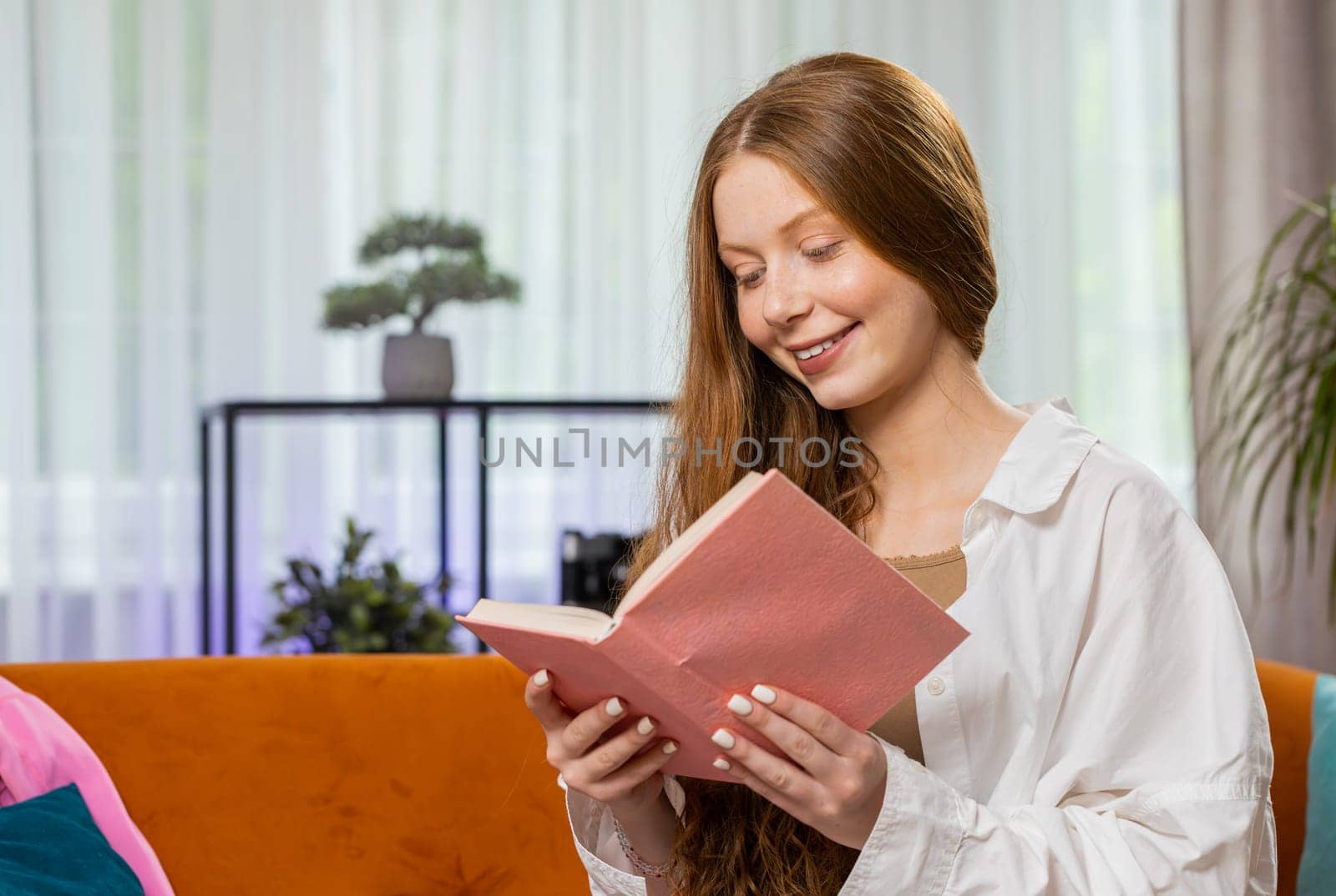 Teenager red hair girl relaxing reading interesting book, turning pages smiling enjoying literature, taking a rest on comfortable couch. Portrait of happy child at home apartment living room on sofa