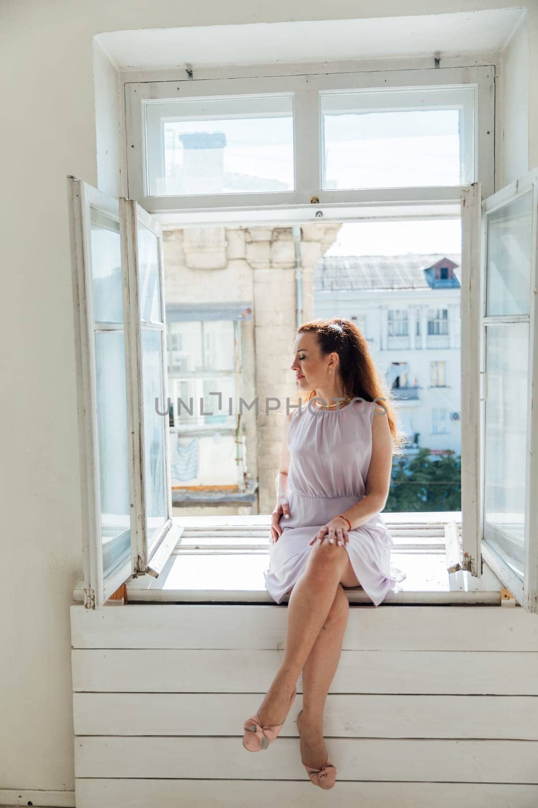 Beautiful brunette woman in dress sitting on windowsill looking at street