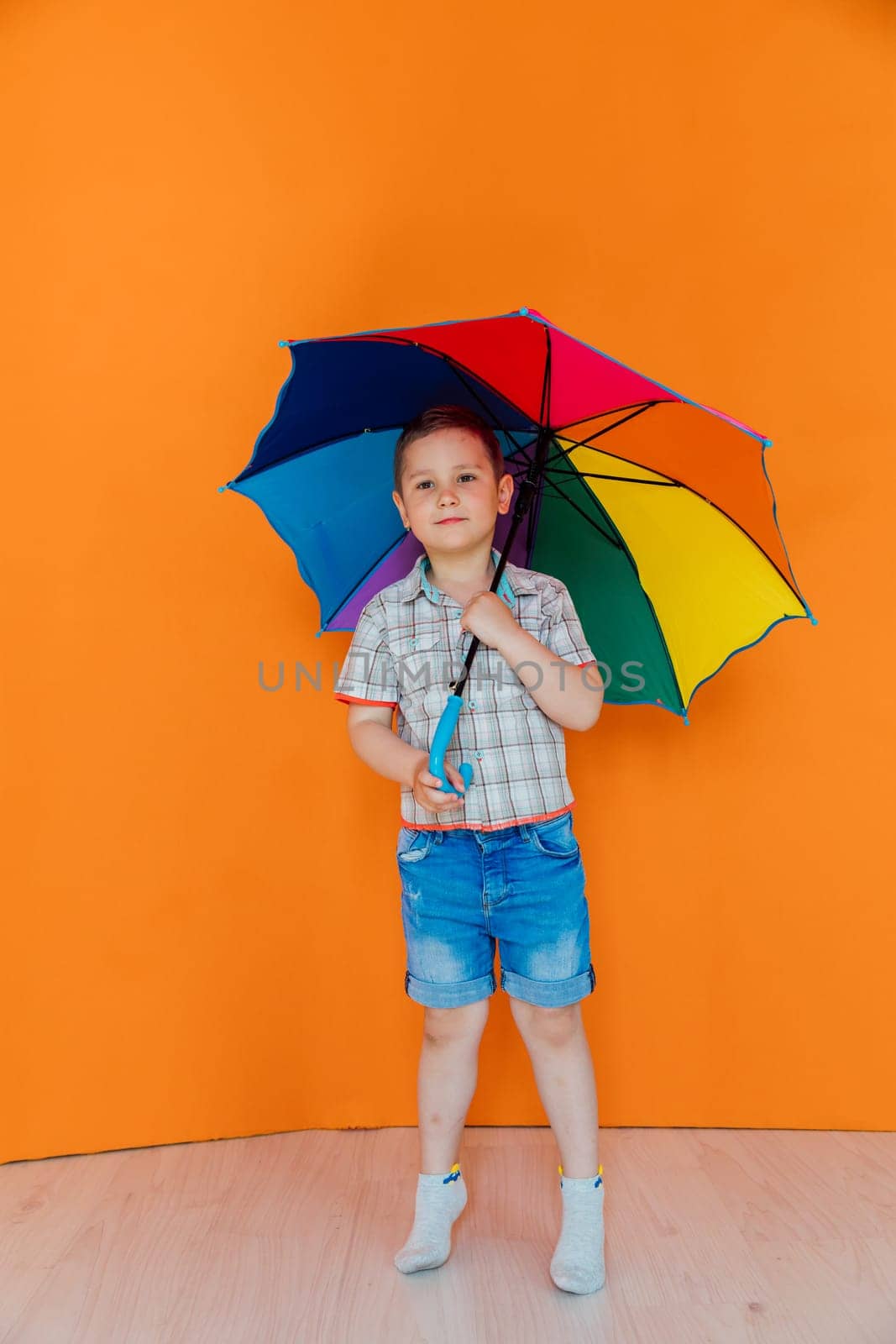 Boy standing under colorful rain umbrella on orange background by Simakov