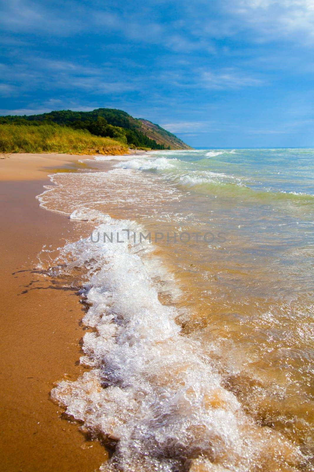 Tranquil beach scene with clear blue sky and vibrant sea. Low perspective captures gentle waves on golden sand. Lush greenery adorns bluff in background.