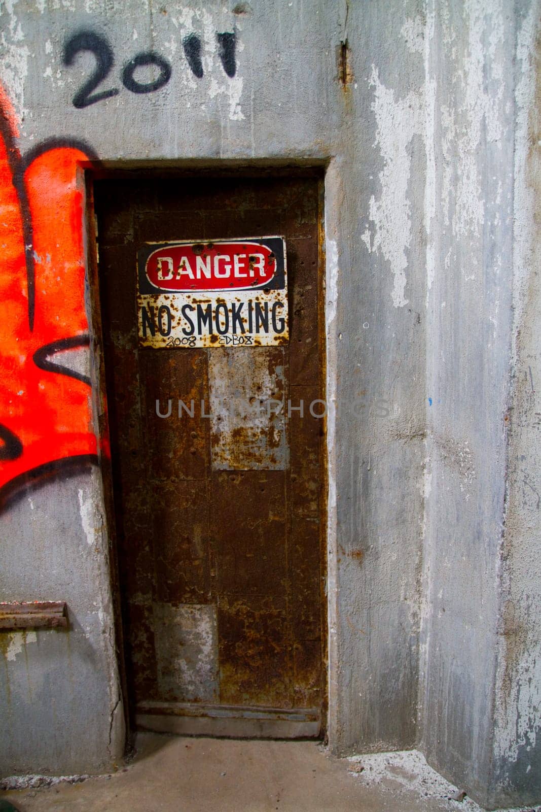 Decaying industrial doorway in East of St. Louis, Illinois. Rusty metal door adorned with weathered 'DANGER NO SMOKING' sign hints at hazardous conditions. Urban decay and cautionary themes.