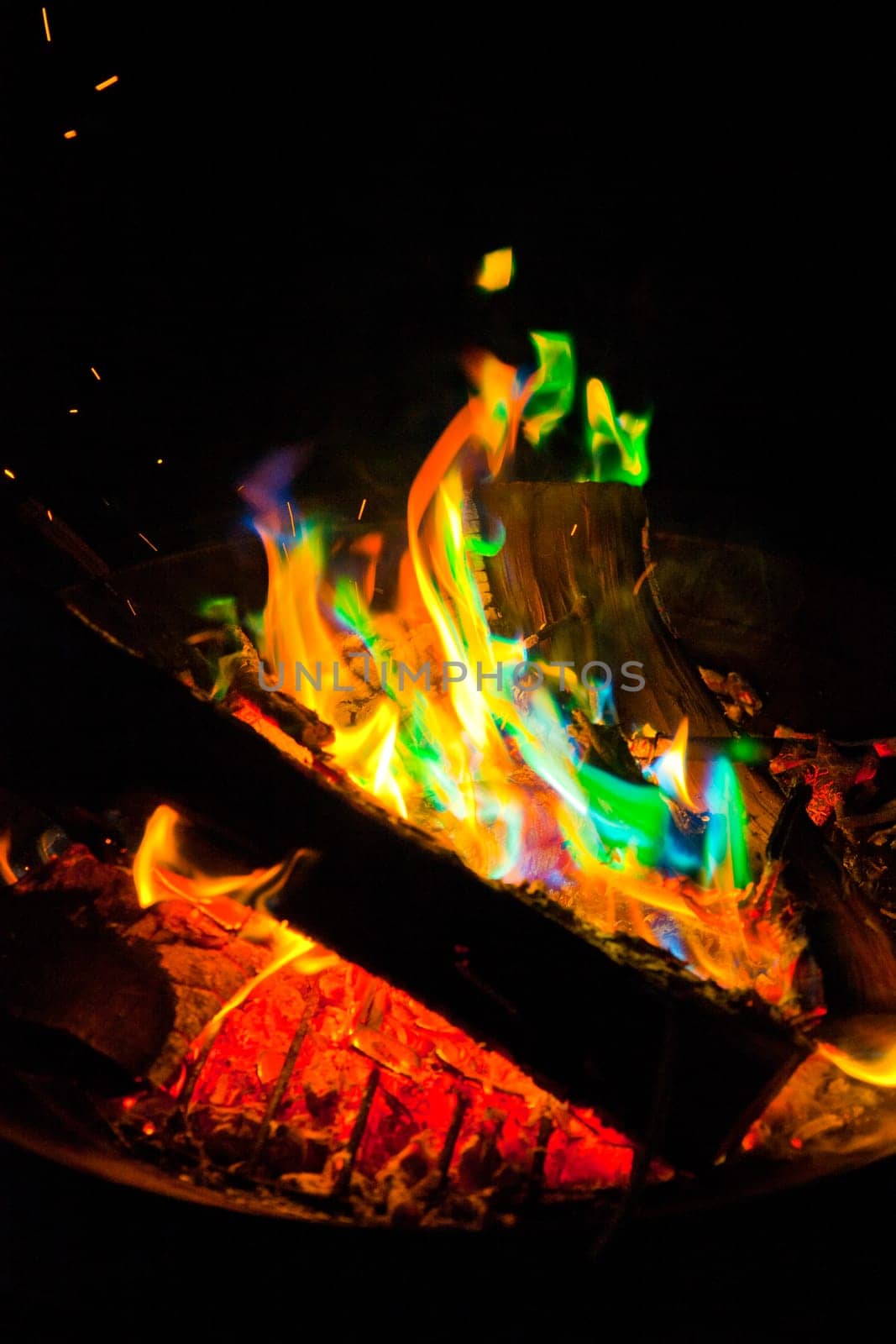 Vibrant and dynamic campfire illuminates the night, with colorful flames flickering in shades of green, blue, orange, and yellow. Sparks dance in the air, adding to the lively atmosphere. Perfect for conveying warmth, energy, and outdoor adventure.