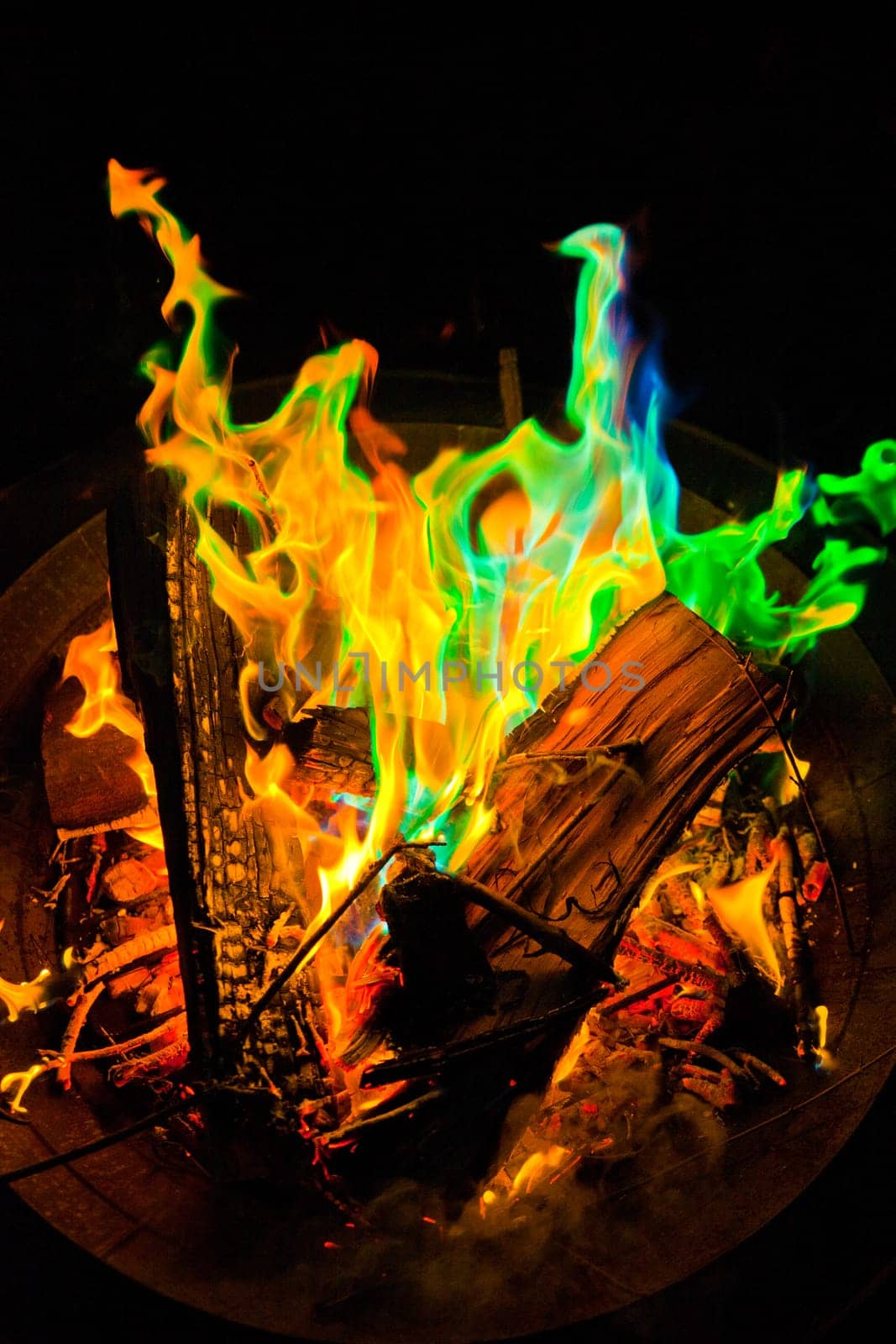 Experience the mesmerizing allure of a vibrant, multi-hued fire in this captivating stock photo. Set within a metal fire pit, the flames dance with green, blue, and traditional orange-yellow tones, creating a mystical and enchanting display.