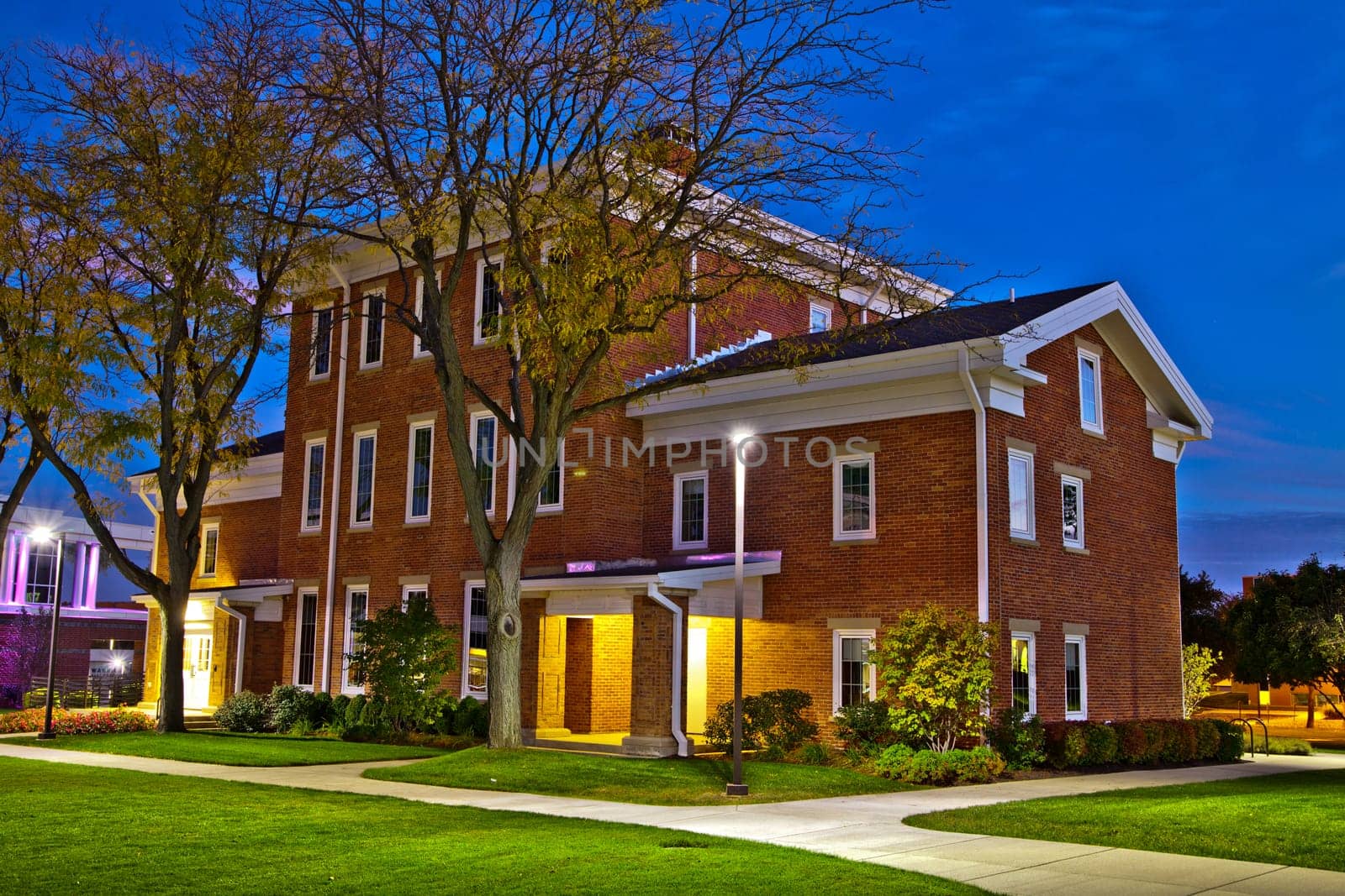 Twilight illuminates a traditional brick building with symmetrical windows and a multi-pitched roof in Fort Wayne, Indiana. The warm artificial light enhances its classic architecture, while the serene urban landscape show early signs of autumn.