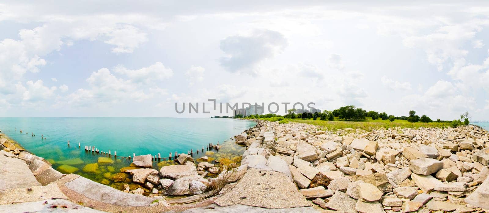 Tranquil lakeside scene with turquoise waters, rugged breakwater, and distant urban skyline - a serene blend of nature and cityscape.