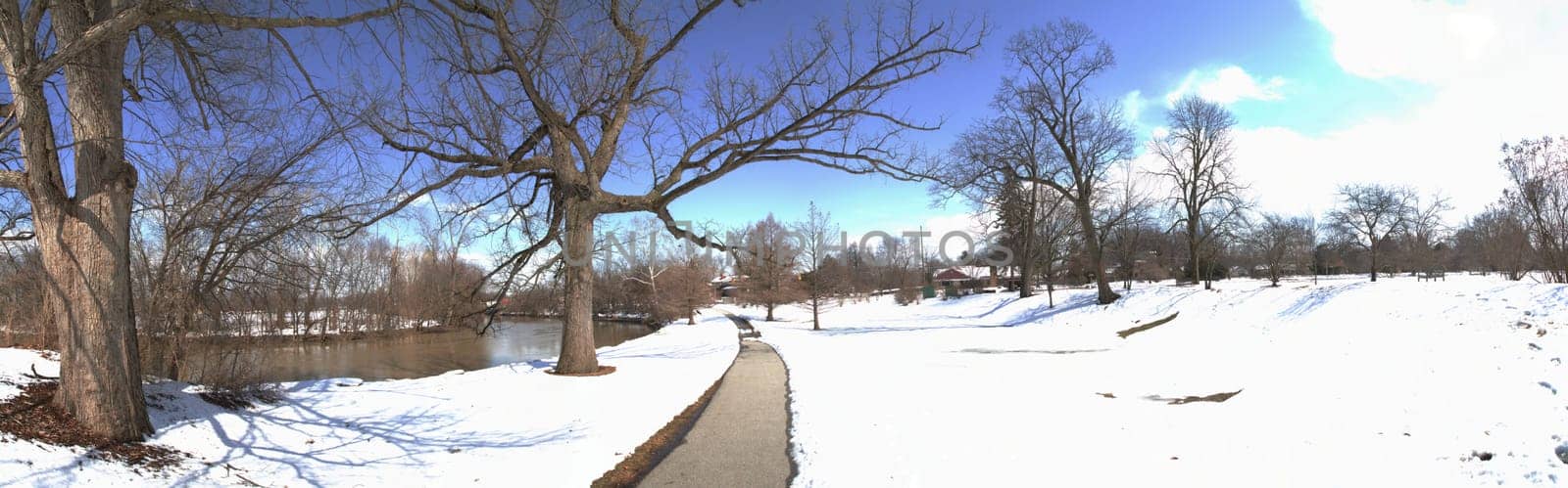 Winter wonderland: A tranquil snow-covered path winds through a serene park, flanked by bare trees and a picturesque river. Perfect for capturing the peaceful solitude and changing seasons.