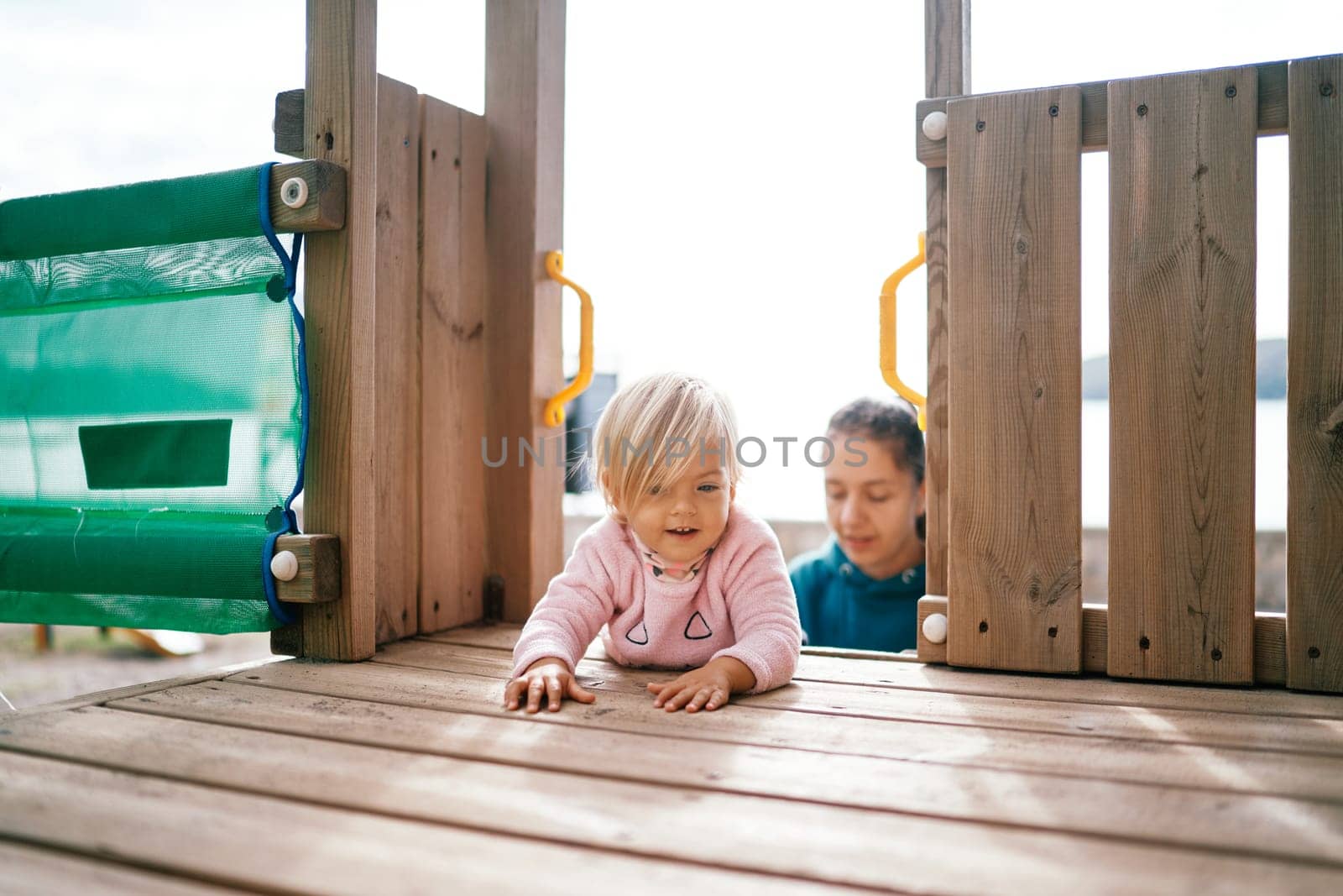 Little girl crawls on her stomach on a wooden deck of a slide against the background of her mother by Nadtochiy