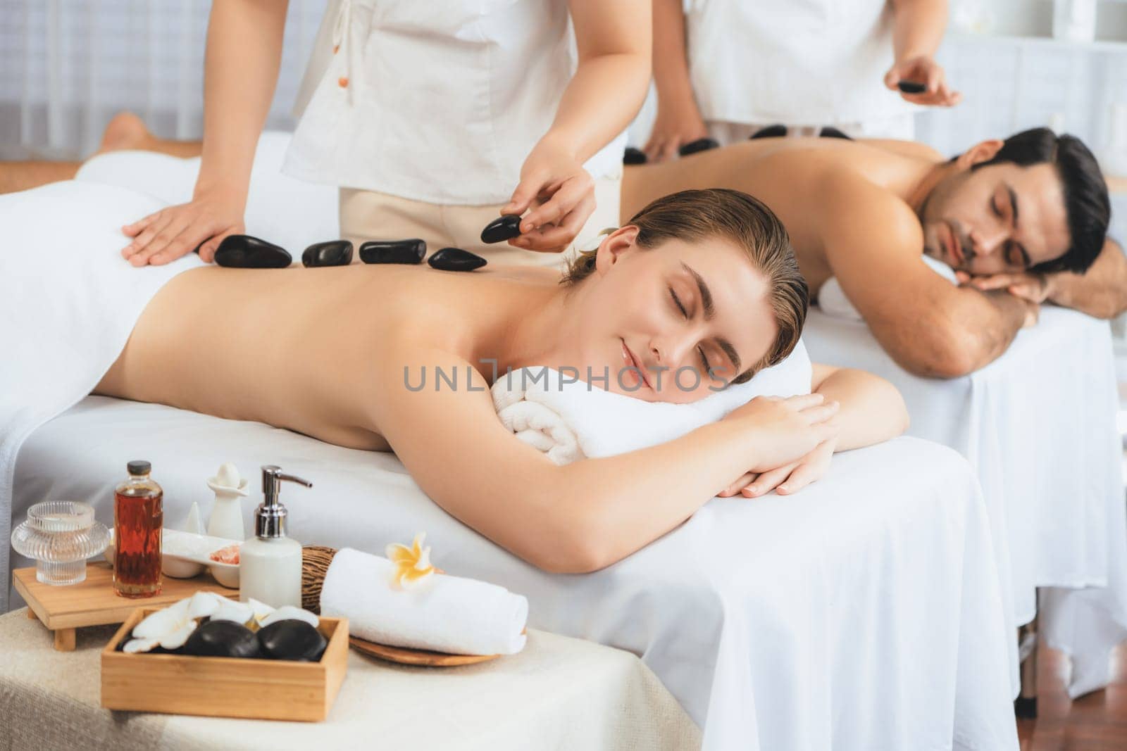 Hot stone massage at spa salon in luxury resort with day light serenity ambient, blissful couple customer enjoying spa basalt stone massage glide over body with soothing warmth. Quiescent