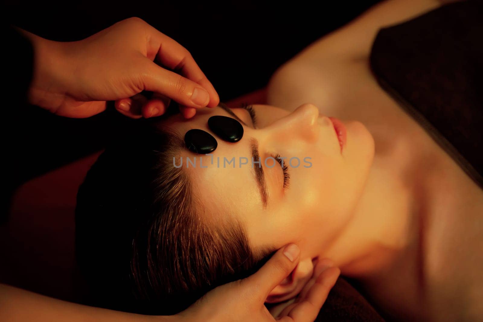 Caucasian woman enjoying relaxing anti-stress head massage with hot stone and pampering facial beauty skin recreation leisure in warm candle light ambient salon spa in resort or hotel. Quiescent