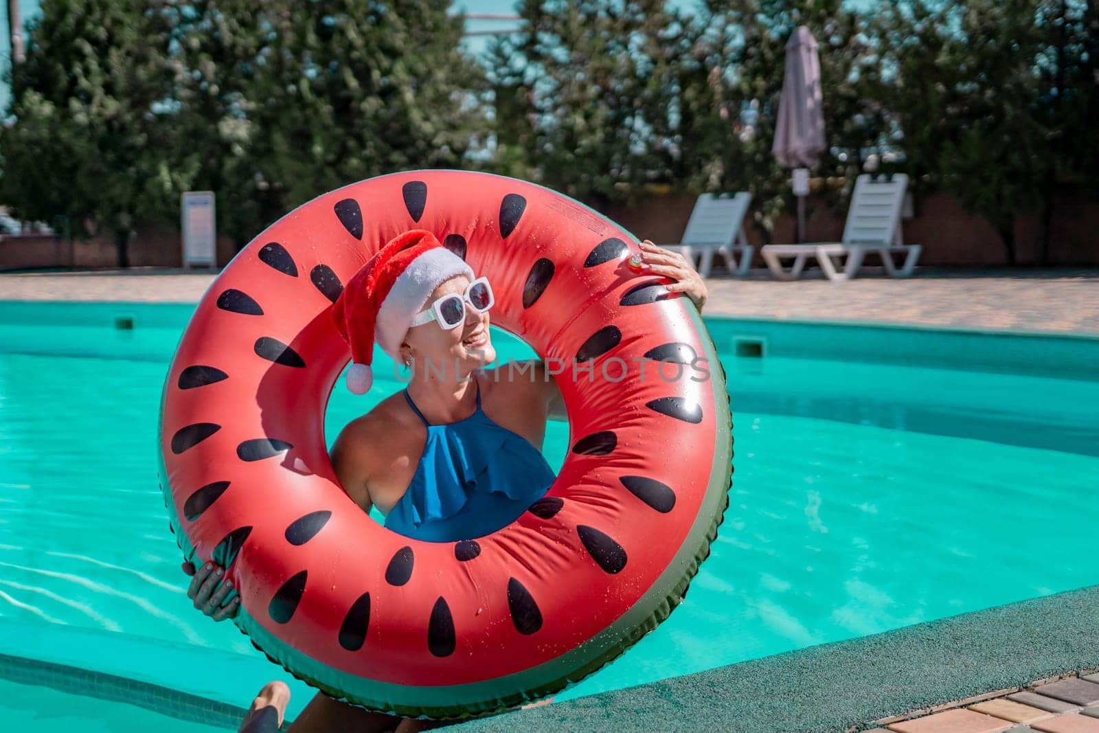 Woman pool Santa hat. A happy woman in a blue bikini, a red and white Santa hat and sunglasses poses near the pool with a glass of champagne standing nearby. Christmas holidays concept. by Matiunina