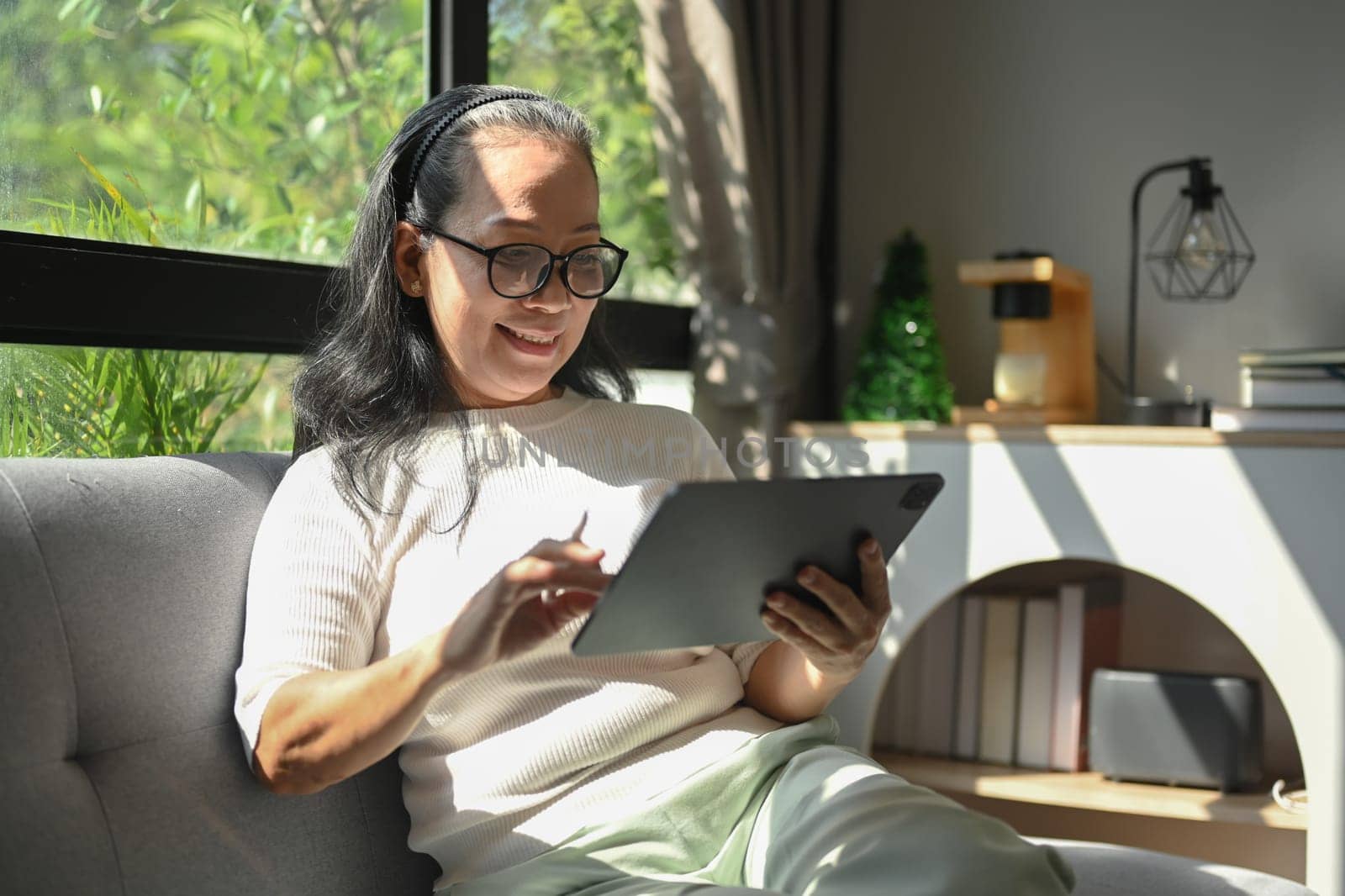 Happy middle age woman using digital tablet on couch in bright living room. by prathanchorruangsak