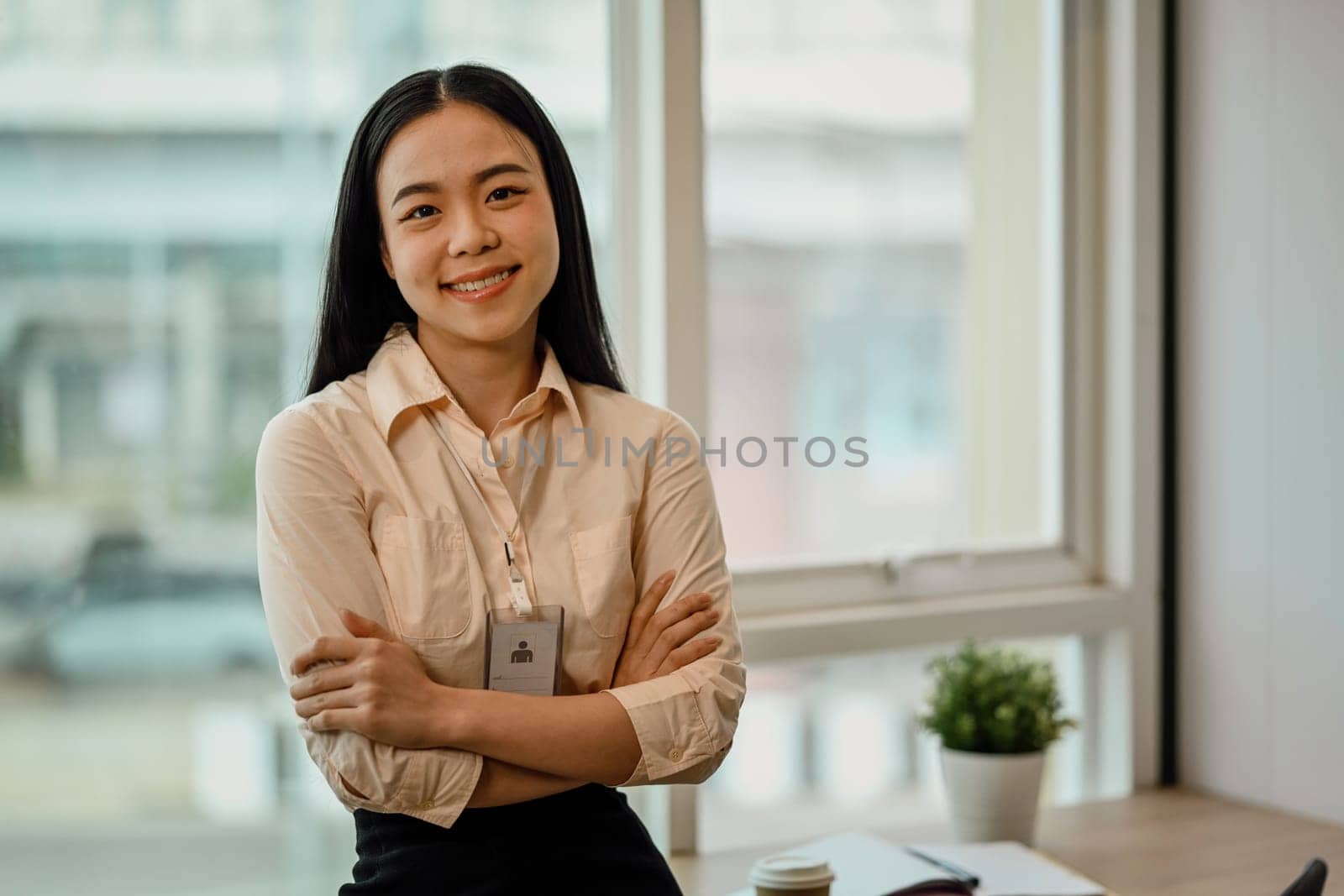 Portrait of a young Asian female employee standing with arms crossed and looks at the camera with a smile