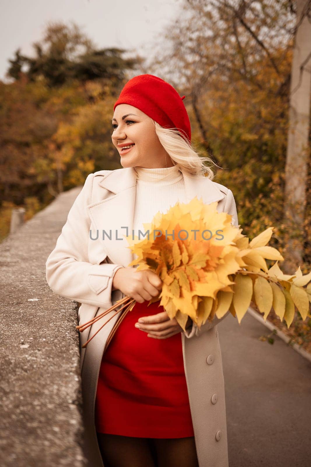 autumn woman in a red beret, a light coat and a red skirt, against the backdrop of an autumn park with yellow leaves in her hands.