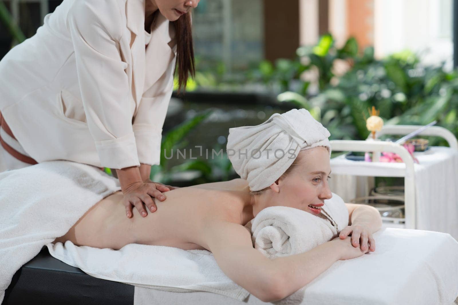 Beautiful young woman received a back massage on a spa bed from masseuse. Attractive female relaxes deeply by skilled hands of the massage therapist. Surrounded with nature. Side view. Tranquility.