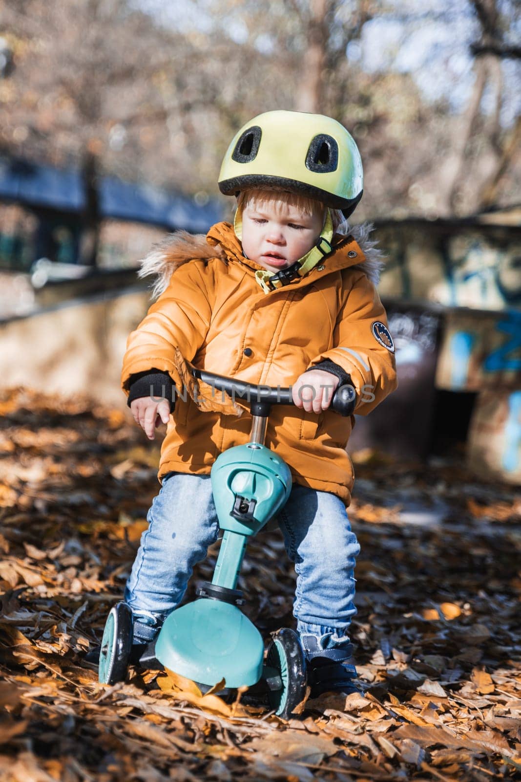 Adorable toddler boy wearing yellow protective helmet riding baby scooter outdoors on autumn day. Kid training balance on mini bike in city park. Fun autumn outdoor activity for small kids