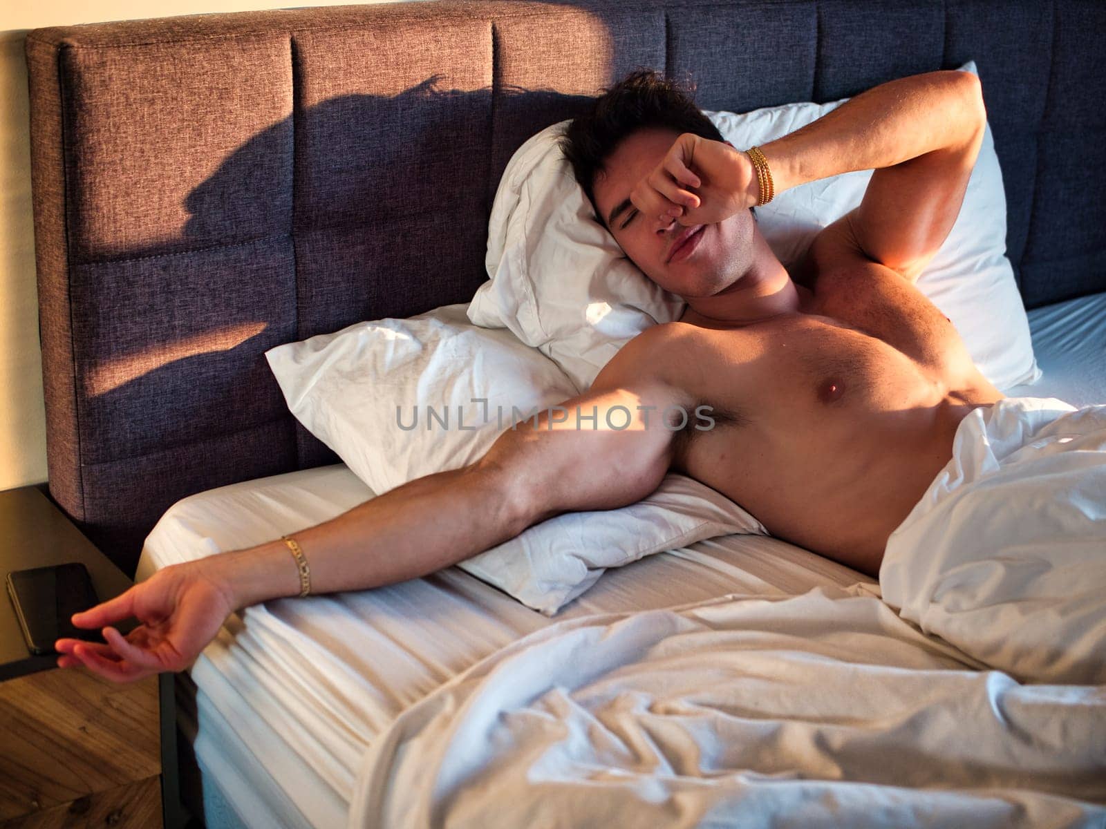 A shirtless man laying on a bed with white sheets, reaching for his cell phone to check his messages