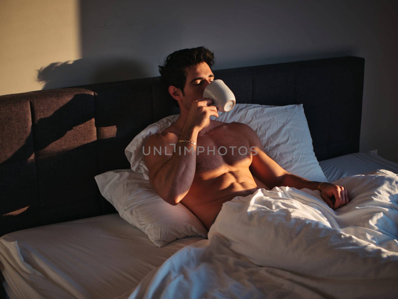A shirtless man laying in bed with a mug, drinking coffee or tea from the cup