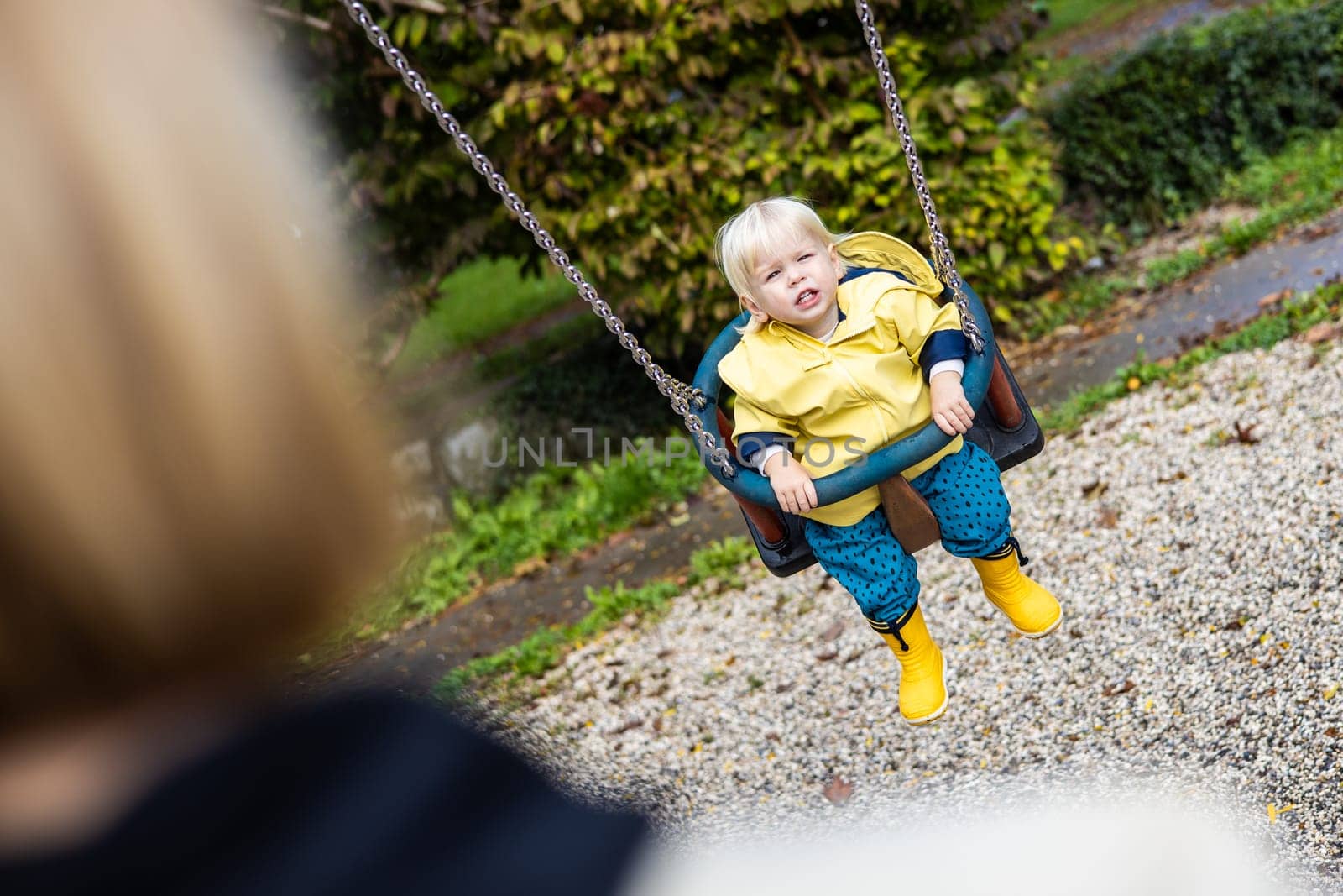 Mother pushing her infant baby boy child wearing yellow rain boots and cape on swing on playground outdoors on cold rainy overcast autumn day in Ljubljana, Slovenia by kasto