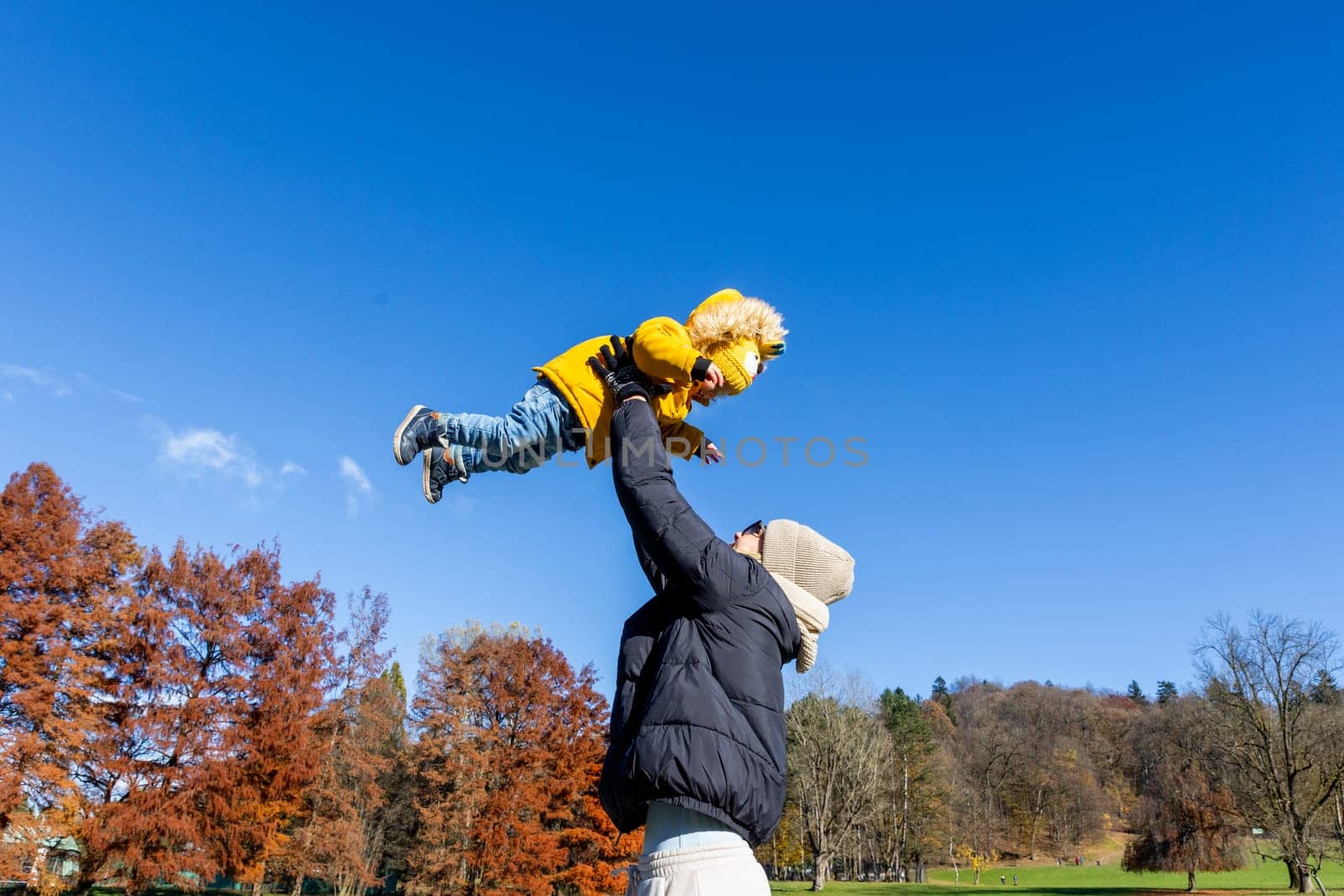 More, more,...mum, that's fun. Happy young mother throws her cute little baby boy up in the air. Mother's Day, Mather and her son baby boy playing and hugging outdoors in nature in fall. by kasto
