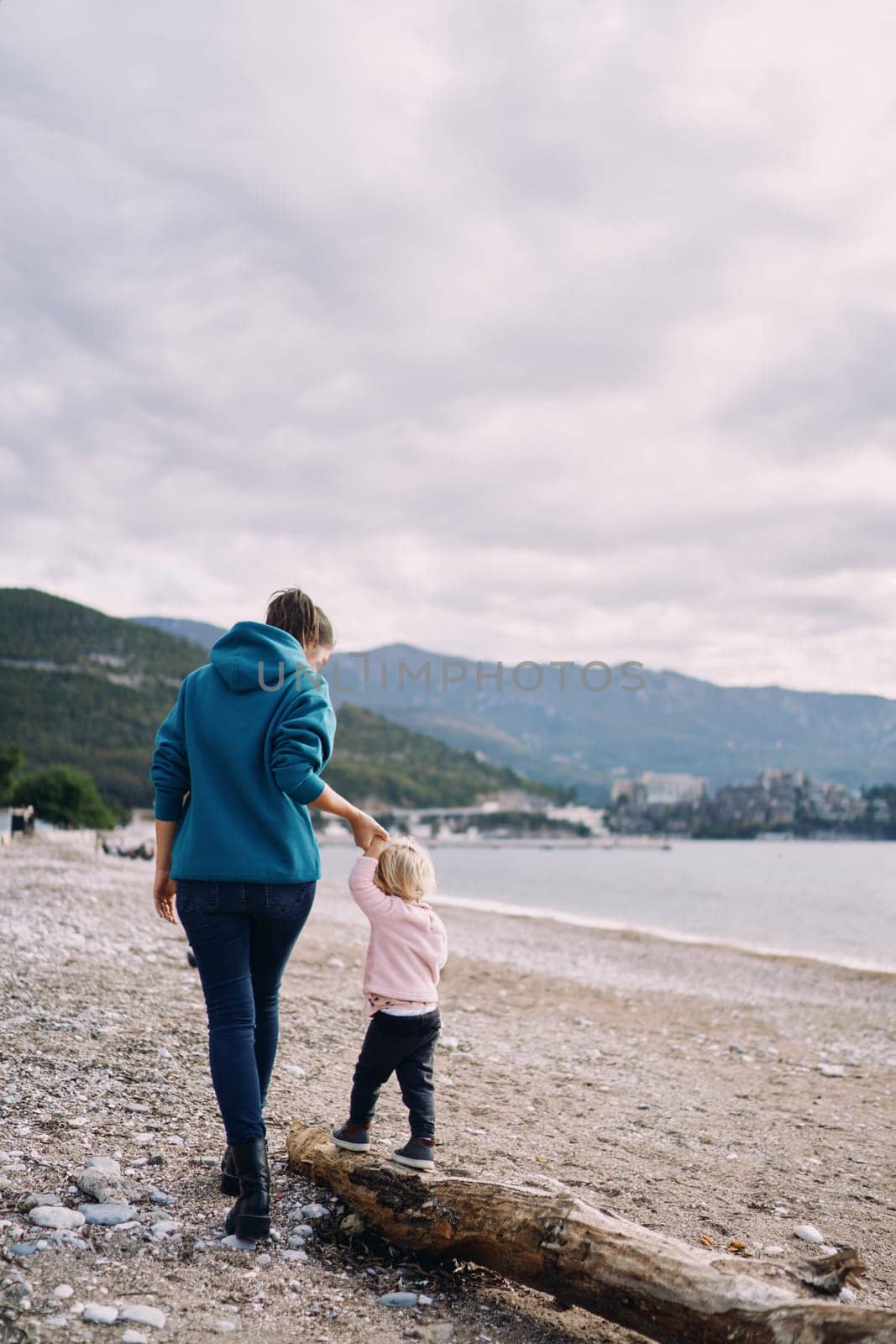 Little girl walks along a driftwood on the beach holding her mother hand. Back view by Nadtochiy