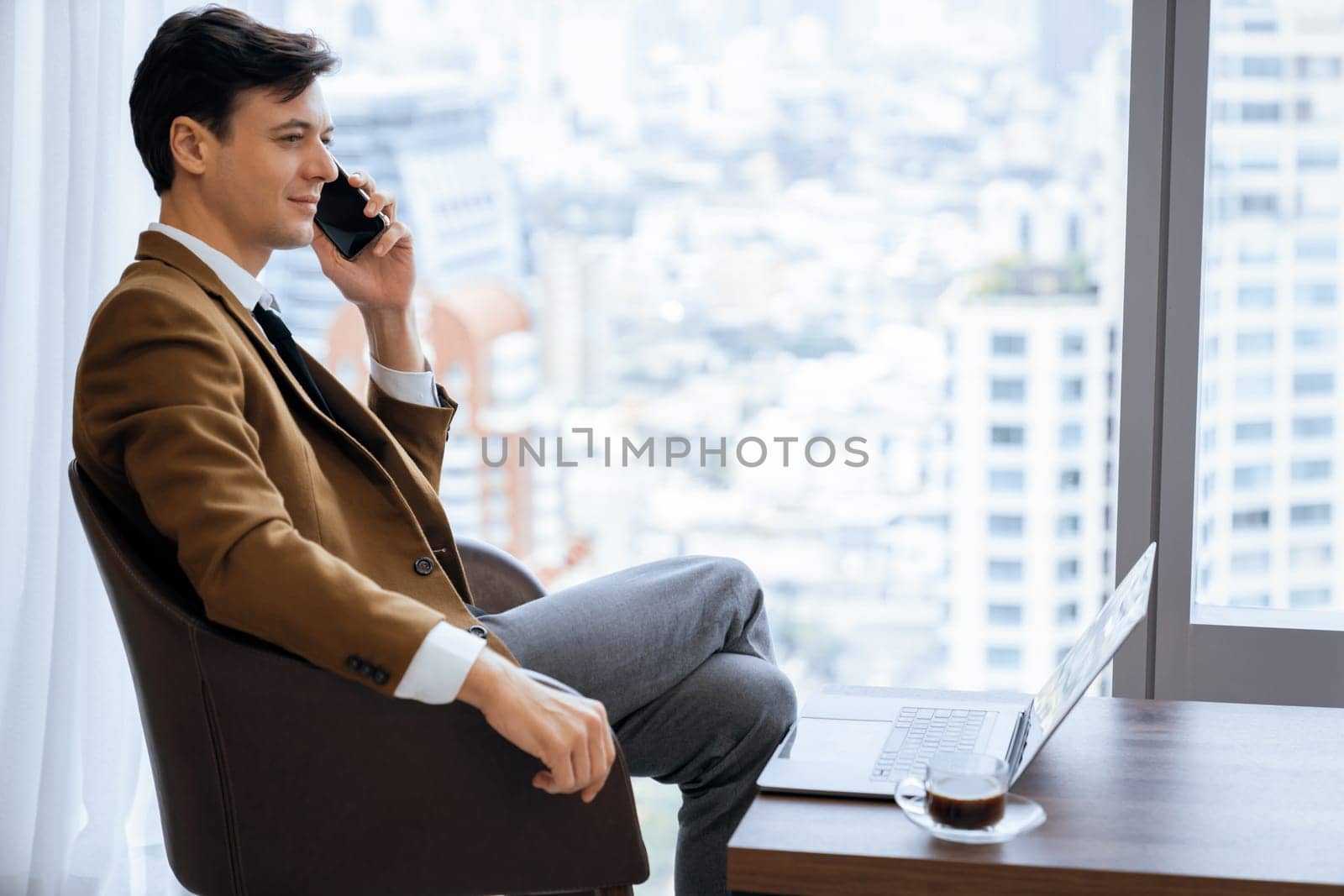 Closeup of handsome businessman making phone call with manager while sitting near window with skyscraper view. Executive manager talking working by using phone and laptop. Look aside. Ornamented.