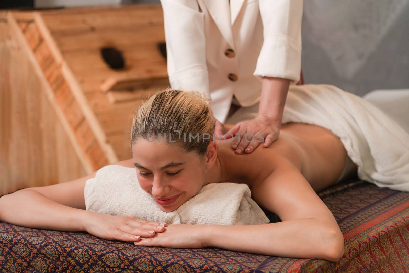 Beautiful young woman lies on spa bed in front of wooden sauna cabinet during having back massage. Attractive women in white towel in warm sauna room. Relaxing and healthy concept Tranquility.