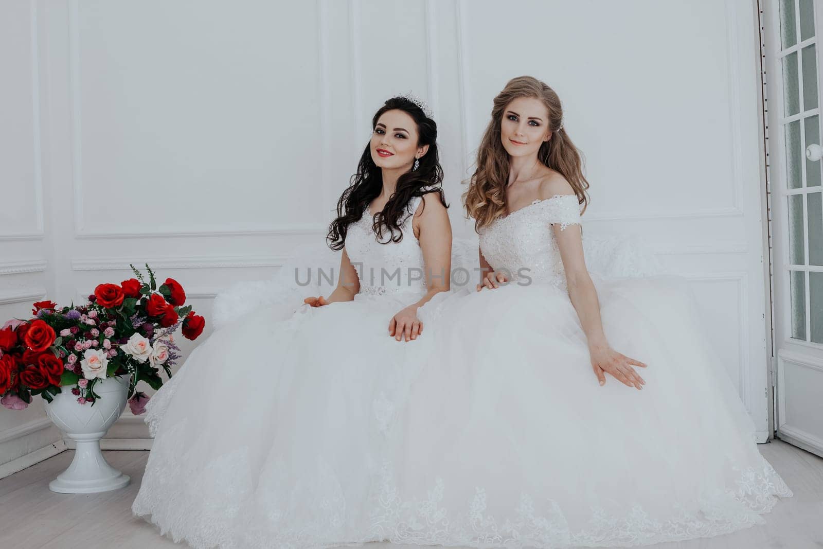 two brides in wedding dresses sit on a white couch in the room