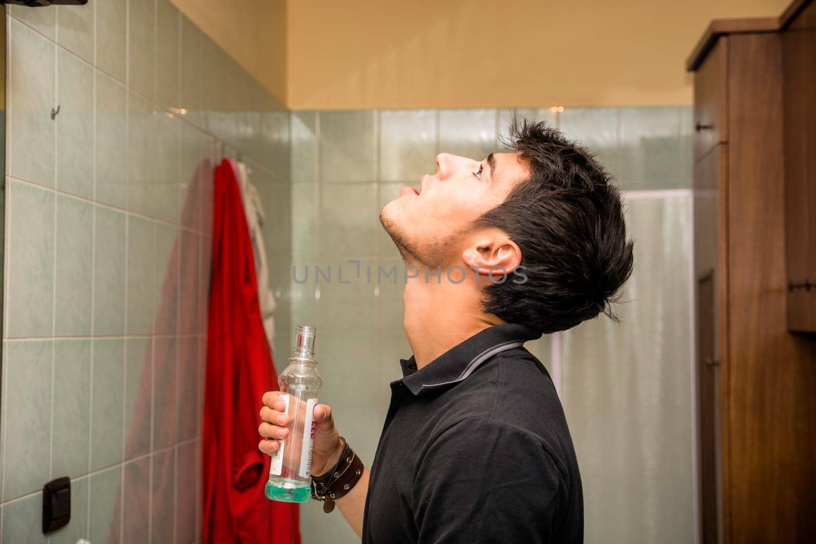 Attractive Young Man with Dark Hair Rinsing with Mouth Wash in Bathroom by artofphoto