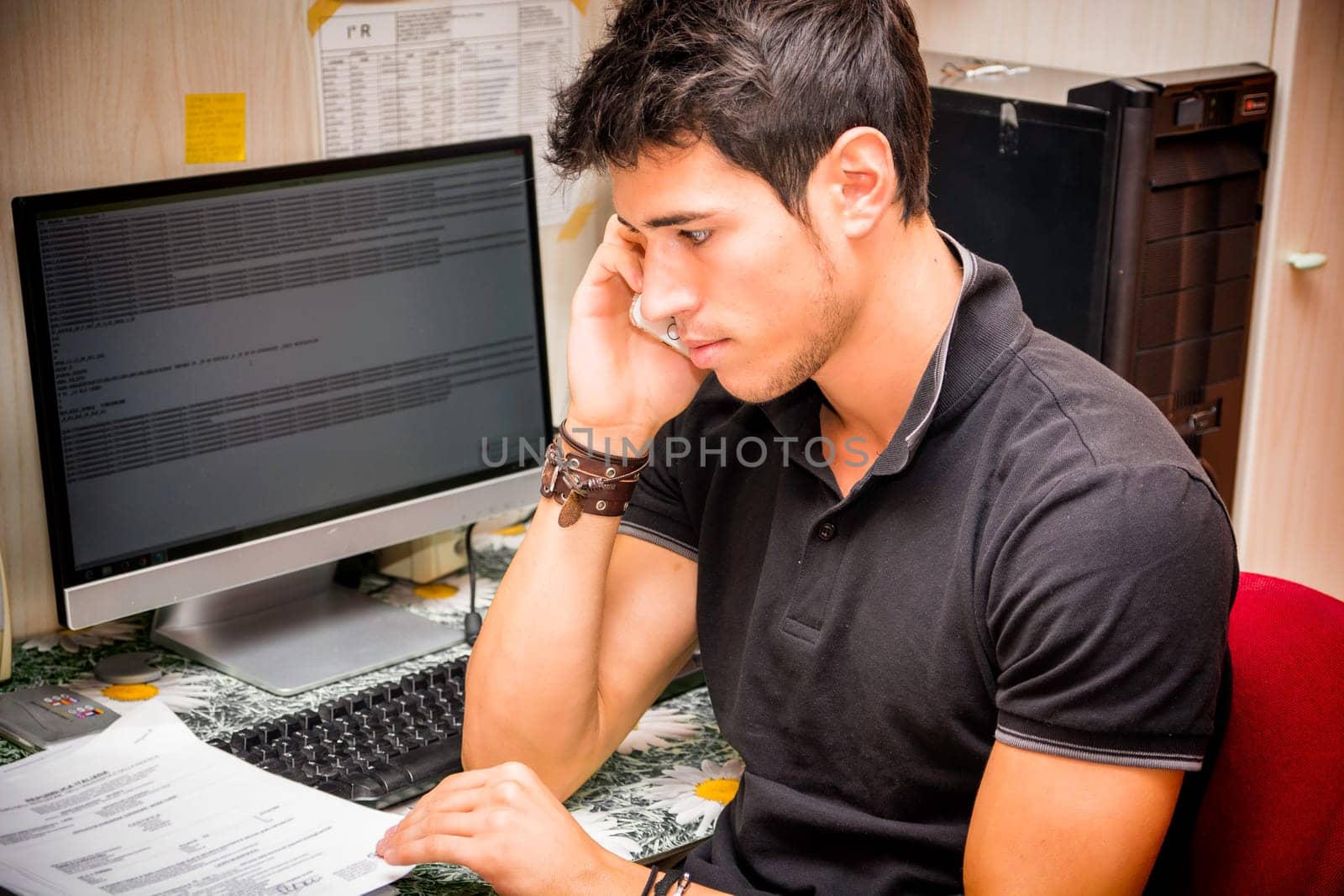 Young Attractive Man Sitting at Computer Desk Talking on Cell Phone by artofphoto