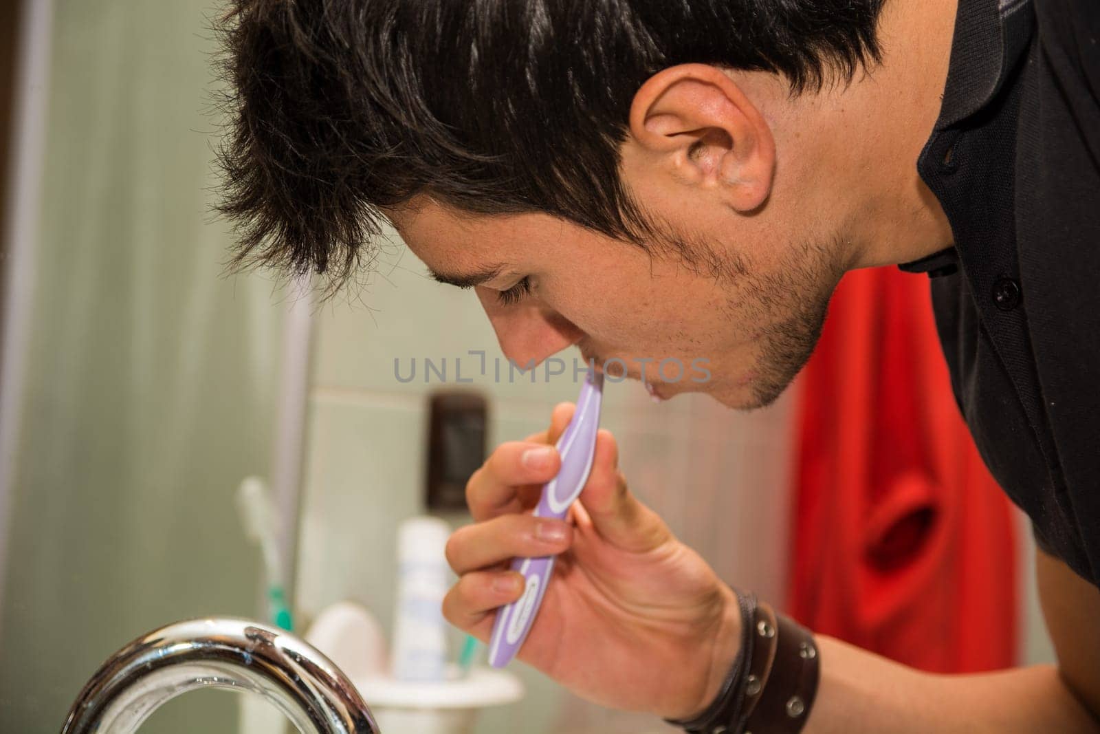 Headshot of attractive young man brushing teeth and cleaning tongue with toothbrush, looking at himself in mirror