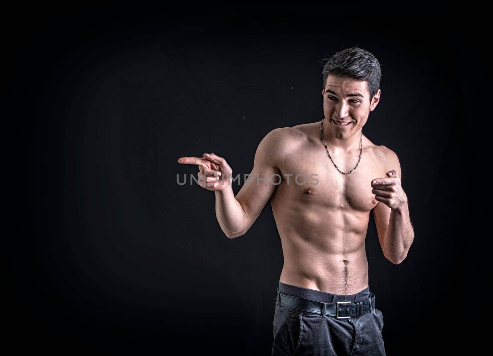 Handsome shirtless young man laughing and pointing finger to a side, on black background