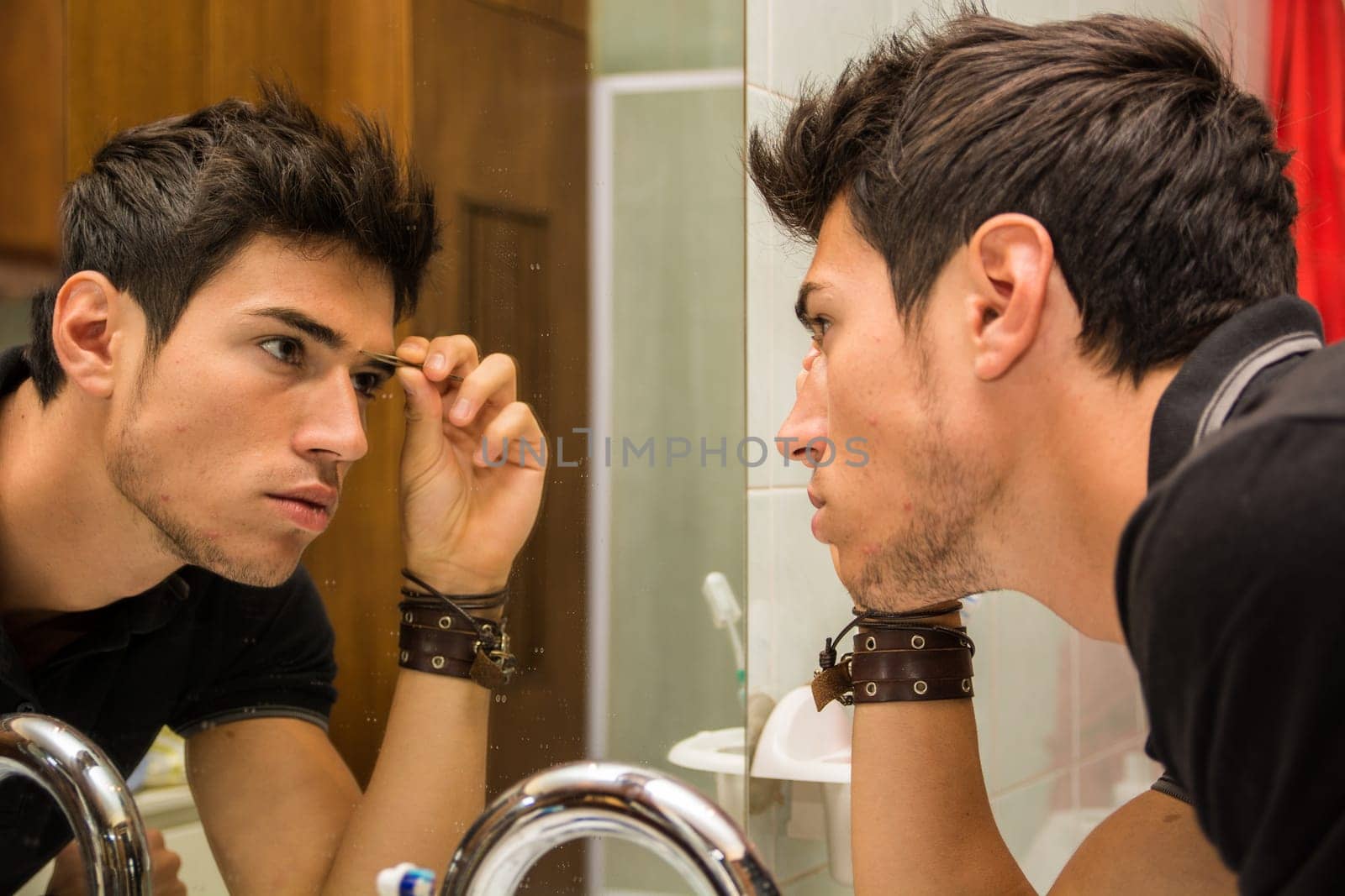 A handsome young man plucking eyebrows in front of a mirror