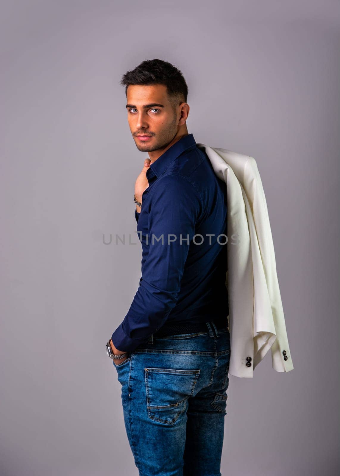 A man in a blue shirt and jeans with a jacket on his back. Photo of a man wearing casual clothing with a jacket slung over his shoulder in studio shot on grey background