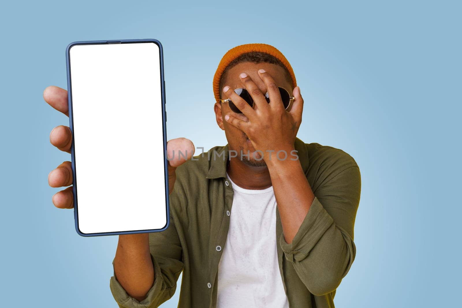 Studio shot of Exited happy young african handsome with sign of facepalm shows mobile phone screen with empty copy space on white display for your text. Man in basic closes isolated on blue background by LipikStockMedia