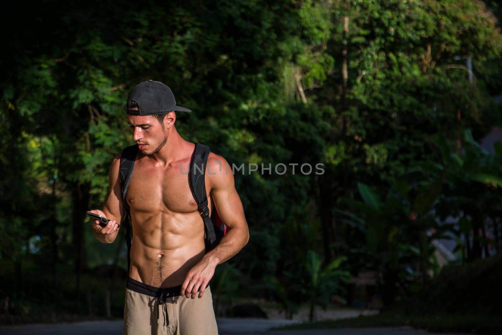 A young fit man without a shirt is walking down the street, looking at his cell phone