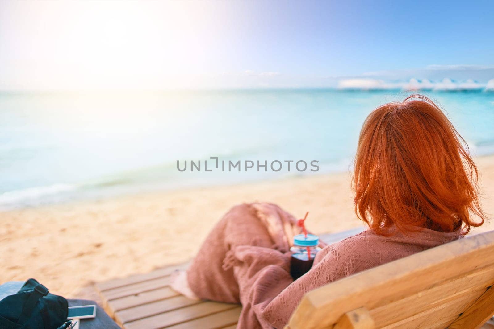 Red-haired girl from behind is lying on chaise longue covered with a blanket. Woman is resting on the sea beach and admiring the ocean landscape. Alcoholic drink in female's hands. Summer holidays.