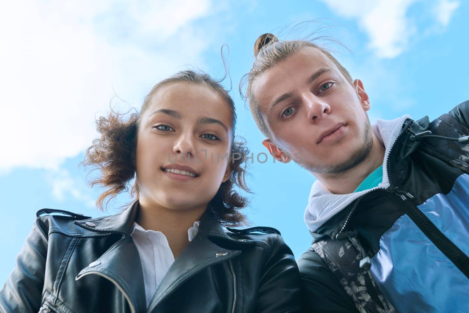 Closeup faces of young teenage guy and girl smiling looking down at camera, blue sky with clouds background. Youth communication leisure friendship lifestyle, college high school students education