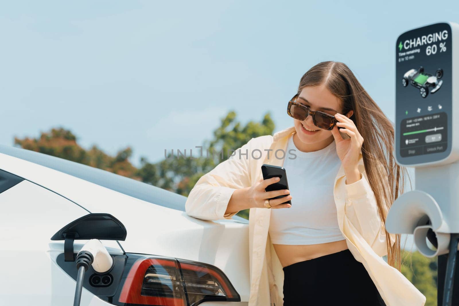 Young woman use smartphone to pay for electricity at public EV car charging station green city park. Modern environmental and sustainable urban lifestyle with EV vehicle. Expedient