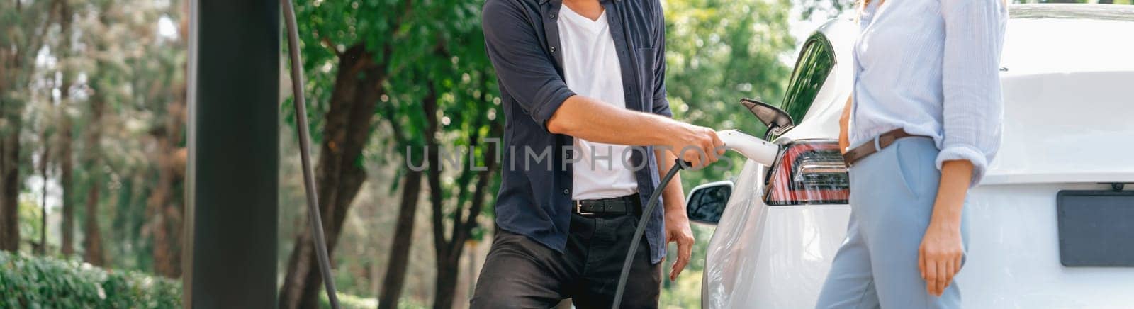 Focused EV car recharging electricity for battery on blurred background of lovey couple ruing their road trip travel by eco friendly electric car in national park and greenery forest on holiday. Exalt